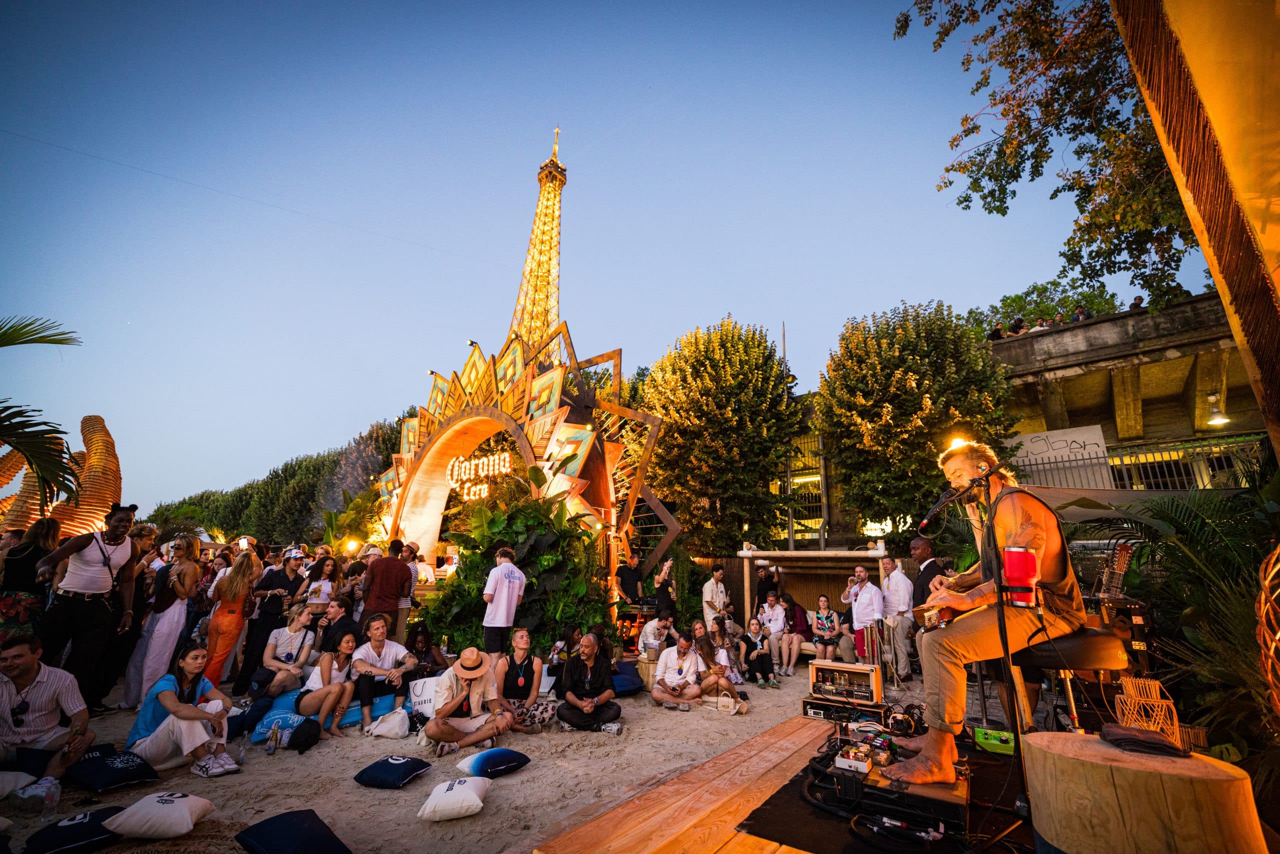 Guests gathered around the Corona Cero Golden Beach stage as a musician performs at dusk beneath the Eiffel Tower
