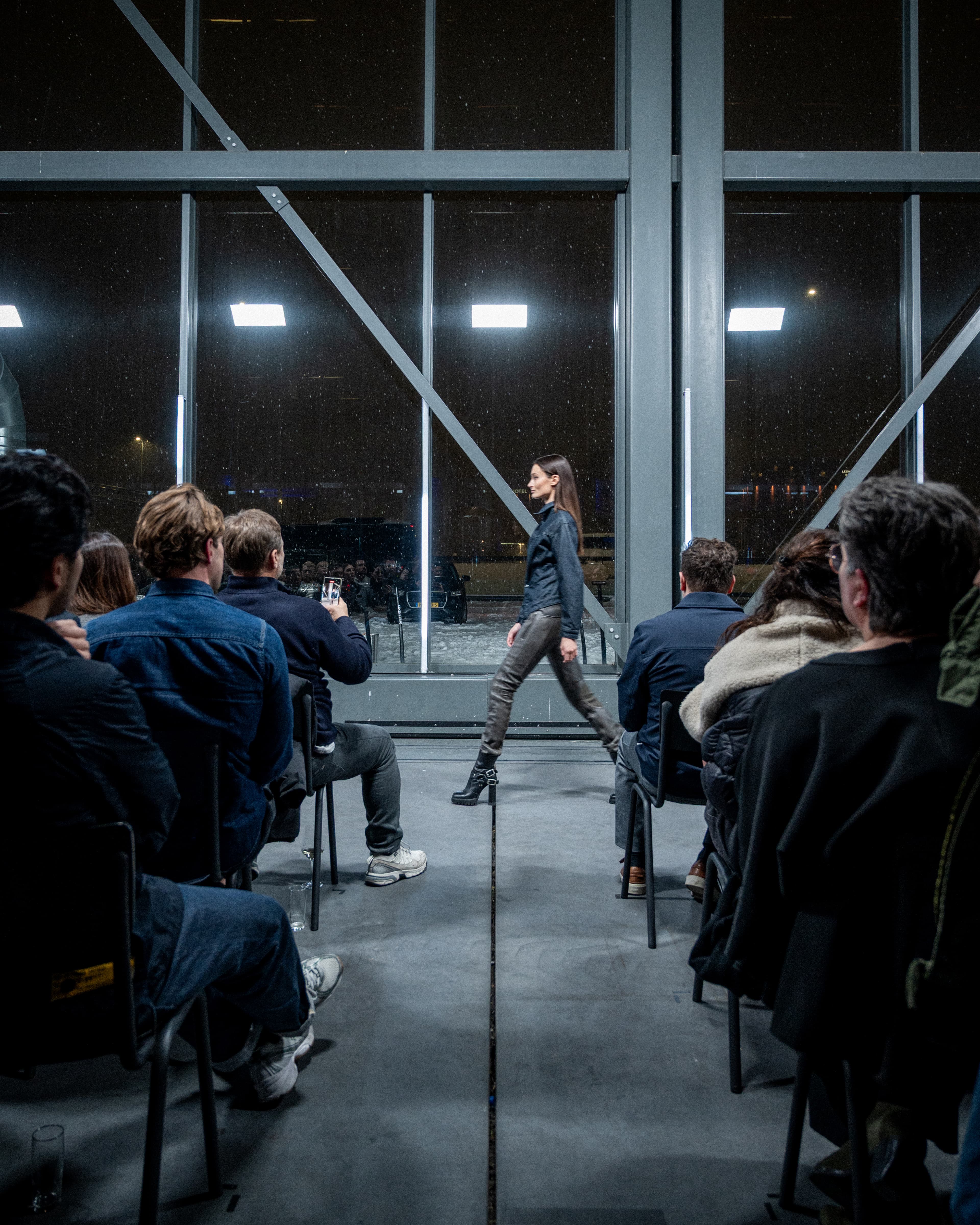 Model walking a concrete runway between seated guests, lit by white strip lights against dark factory windows