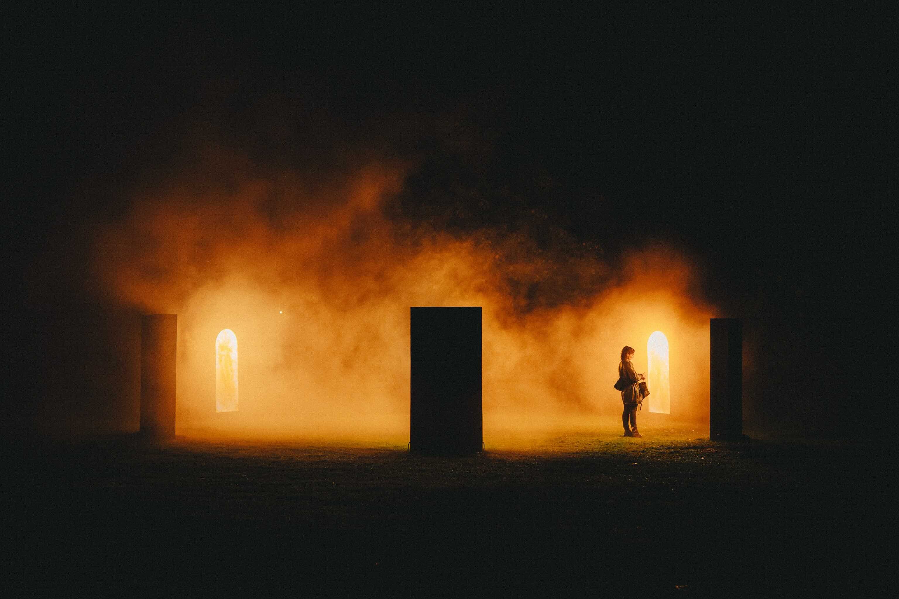 Een eenzame bezoeker in oranje verlichte mist tussen donkere zuilen en twee gloeiende boogvormige deuropeningen in een park bij nacht