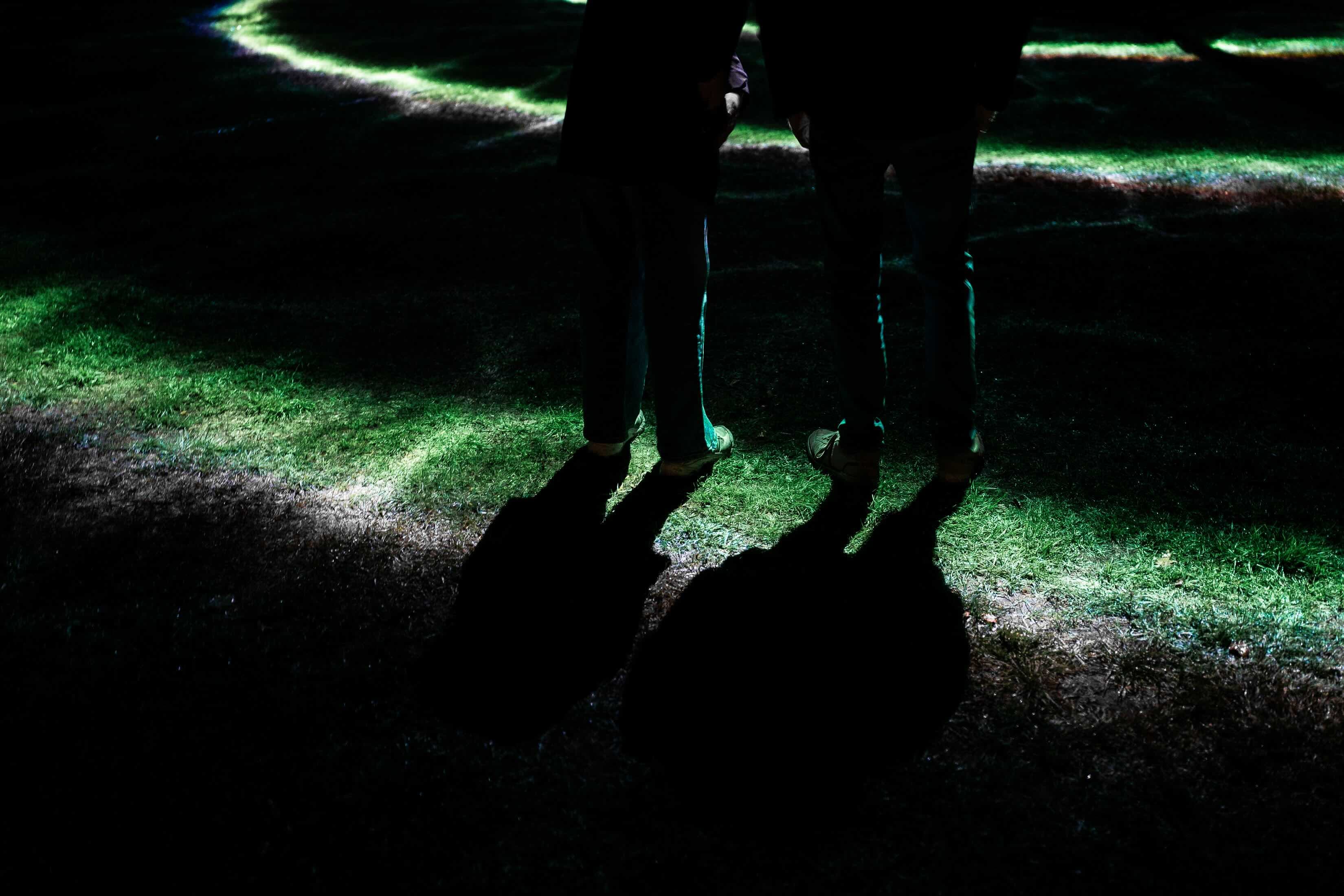 Benen en lange schaduwen van twee bezoekers op gras met strepen groen geprojecteerd licht