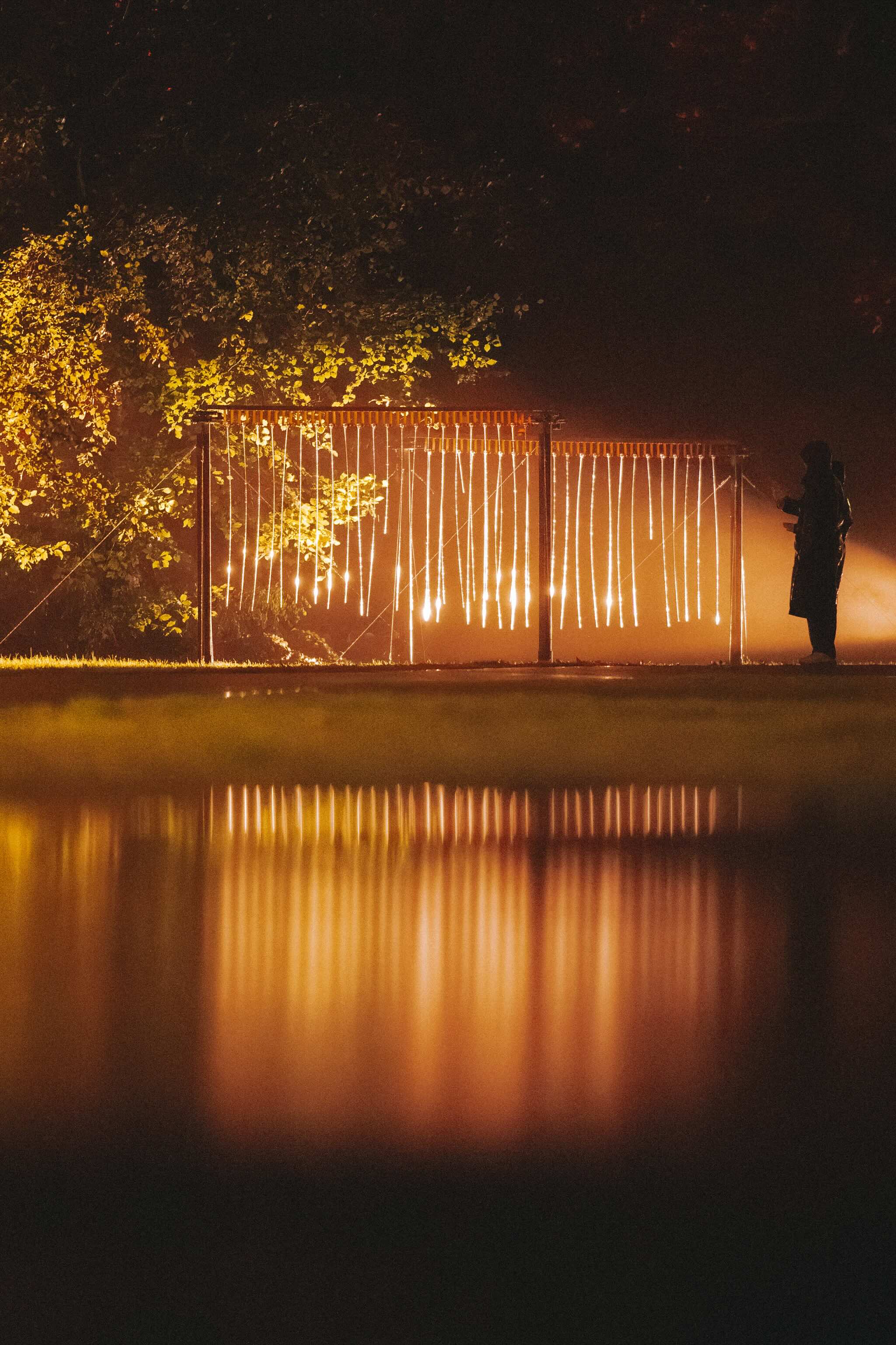 Frame met hangende strengen warm licht naast bomen, weerspiegeld in stil water, met het silhouet van een bezoeker aan de rand