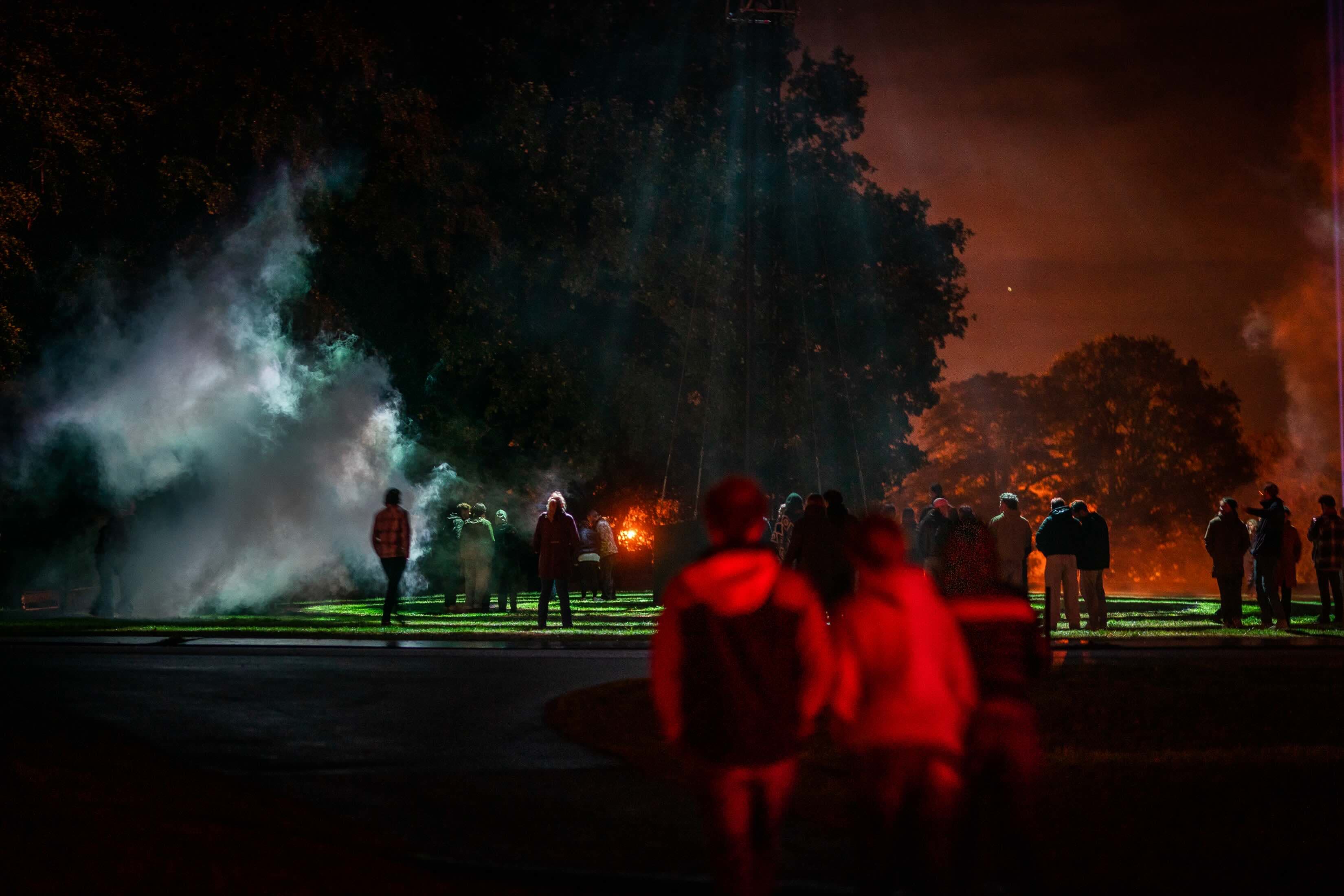 Bezoekers lopen over een groen verlicht gazon naar opwaaiende rook en een rode gloed onder hoge bomen