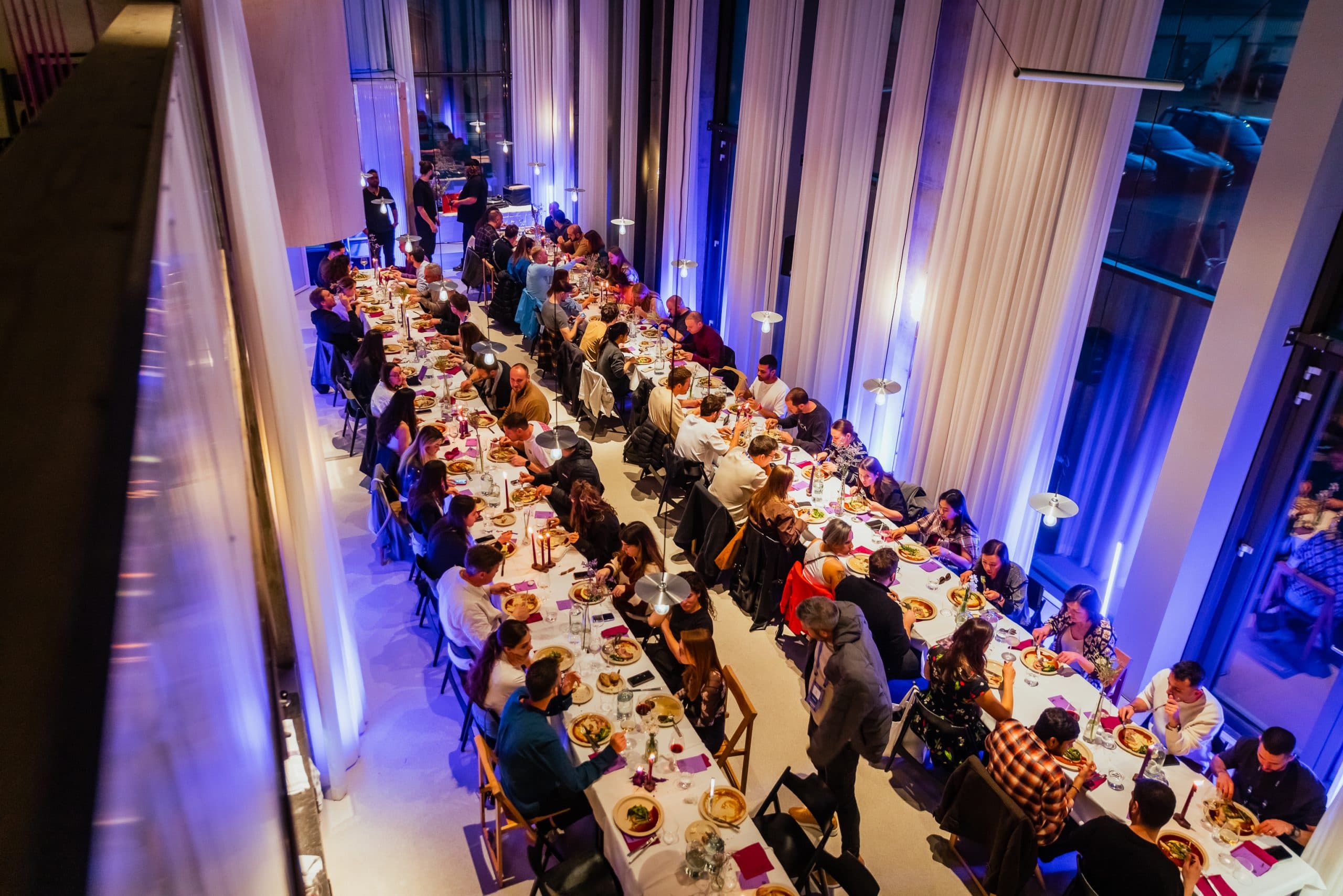 A long banquet table seen from above, lit blue against tall glass walls