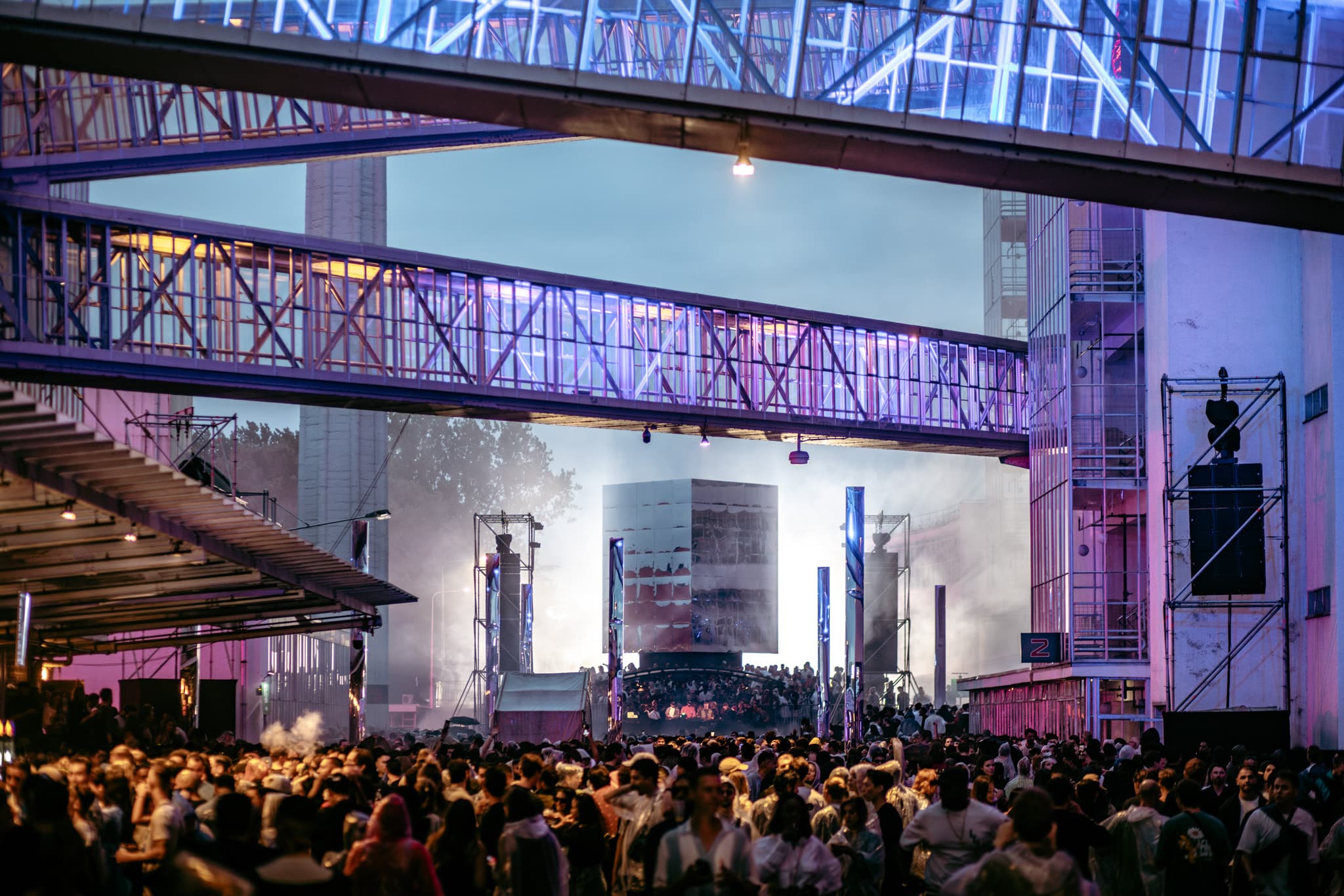 Audio Obscura at Van Nelle Fabriek: a crowd, mirrored stage and illuminated industrial skybridges at dusk