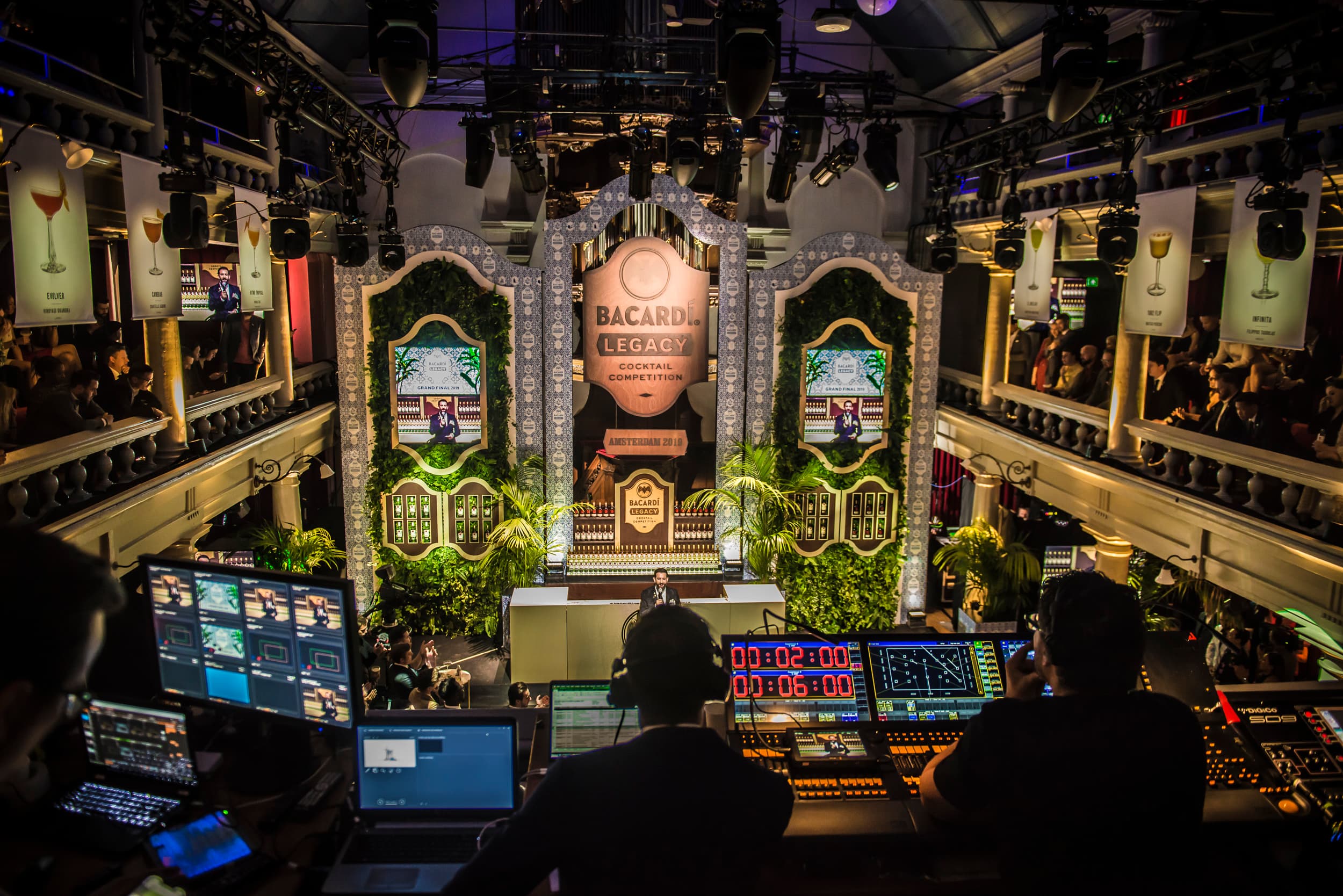 The view over a front-of-house desk: operators at consoles and countdown clocks facing a lit competition set