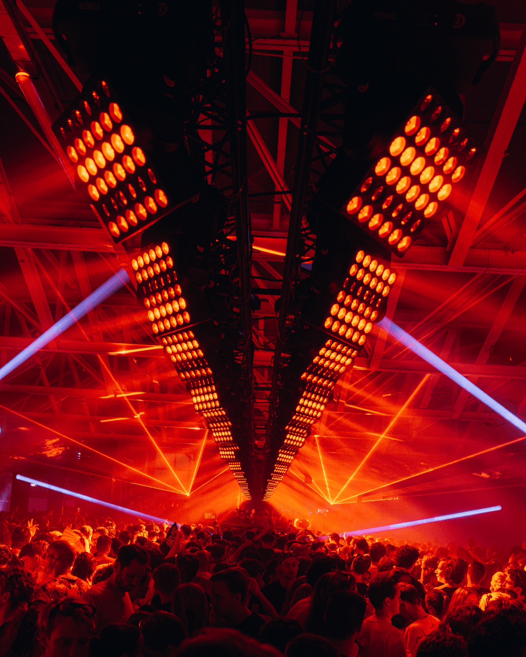 Two converging arrays of orange lights above a dense crowd, with red laser lines cutting through the haze
