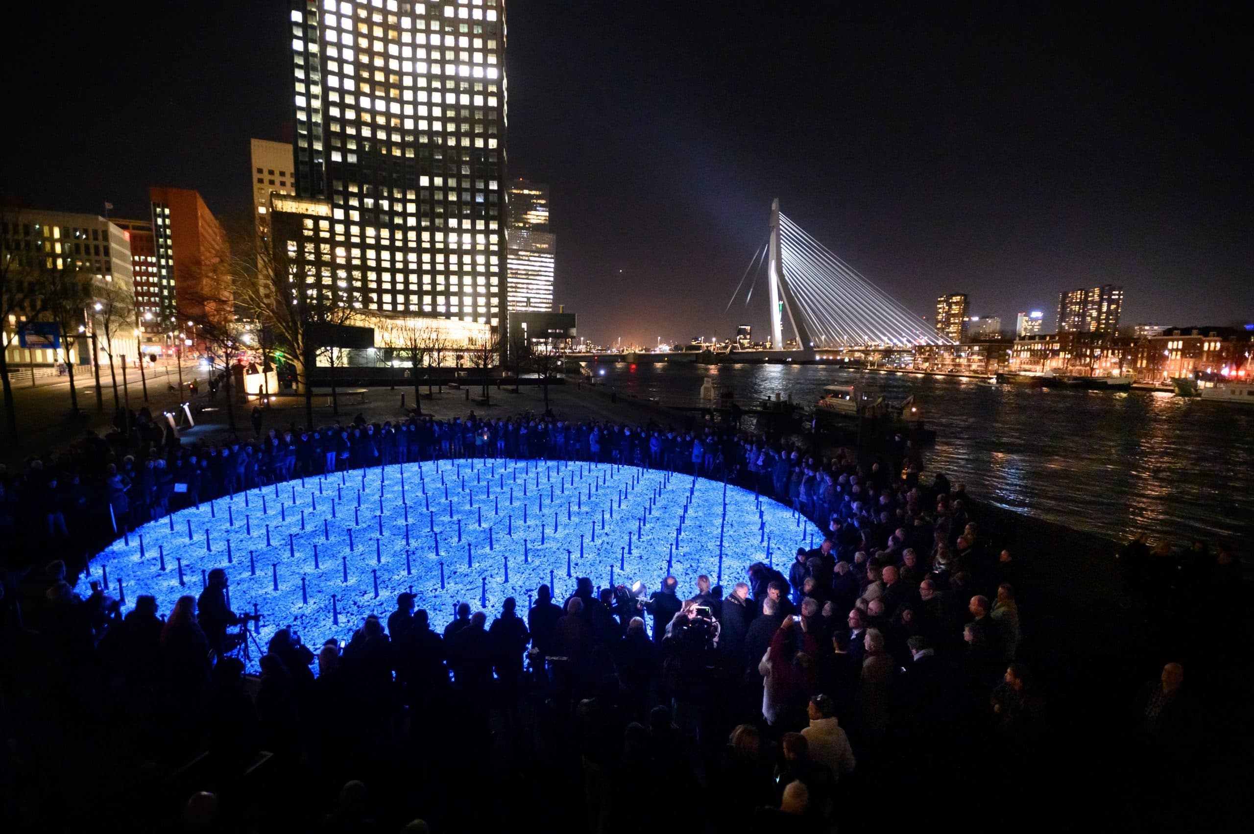 Crowd gathered around the circular blue Levenslicht installation beside the illuminated Rotterdam skyline at night