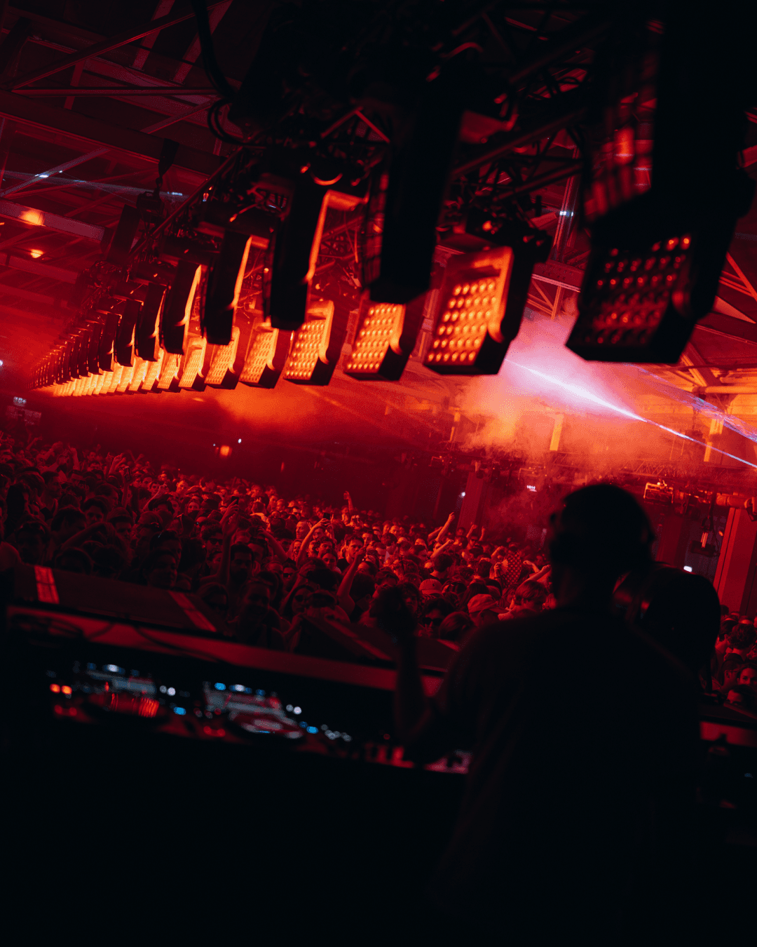 Row of red LED panels above a packed dance floor, seen from behind the DJ booth through haze