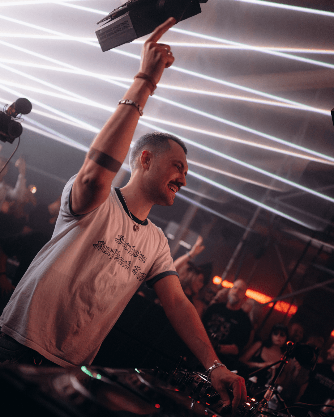 DJ raising one arm behind the decks as pale beams of light cross the ceiling above him